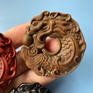 K&ouml;nnte beinhalten: Zwei aufwendig geschnitzte Drachen-Anh&auml;nger aus Holz. Einer ist dunkel rotbraun und der andere ist heller braun mit einem detaillierteren Design.