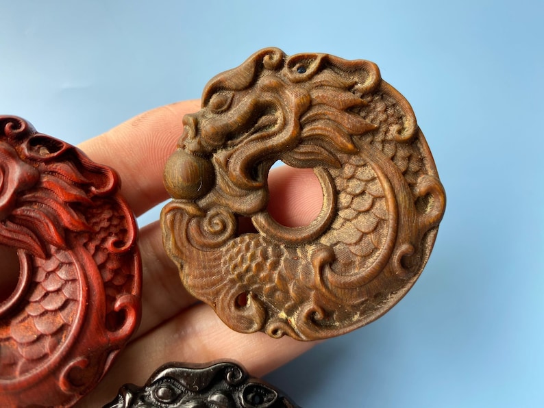 K&ouml;nnte beinhalten: Zwei aufwendig geschnitzte Drachen-Anh&auml;nger aus Holz. Einer ist dunkel rotbraun und der andere ist heller braun mit einem detaillierteren Design.