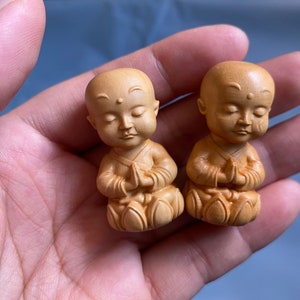 May include: Two small, wooden monk figurines with serene expressions and hands clasped in prayer. The figures are carved in a light brown color, with detailed robes and lotus-like bases. The background is a soft blue.