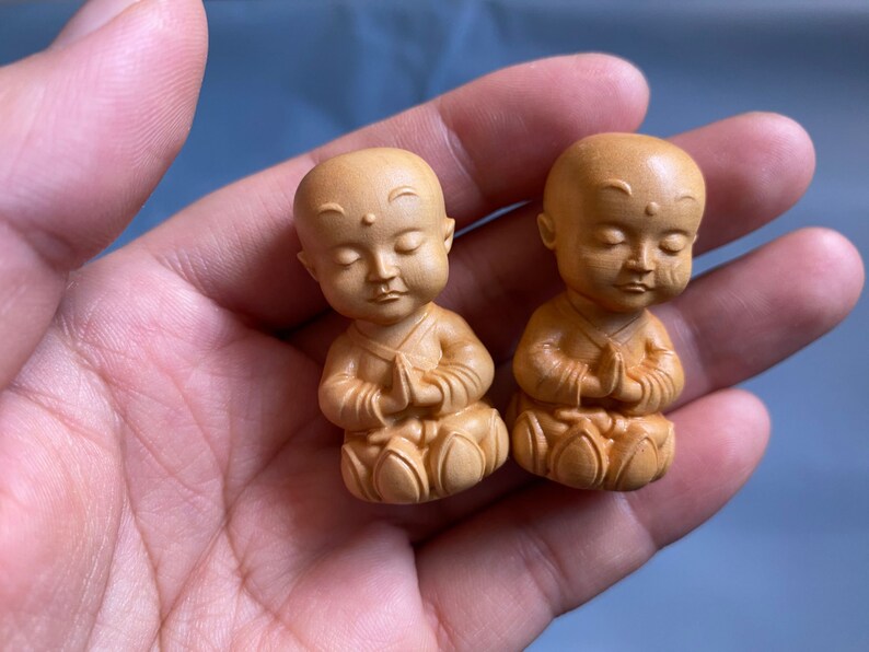 May include: Two small, wooden monk figurines with serene expressions and hands clasped in prayer. The figures are carved in a light brown color, with detailed robes and lotus-like bases. The background is a soft blue.