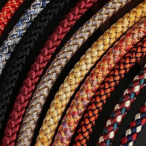 May include: A selection of braided cords in a range of colours, including red, blue, black, gold, and orange. The cords are arranged diagonally, displaying their intricate woven patterns and textures against a dark background.