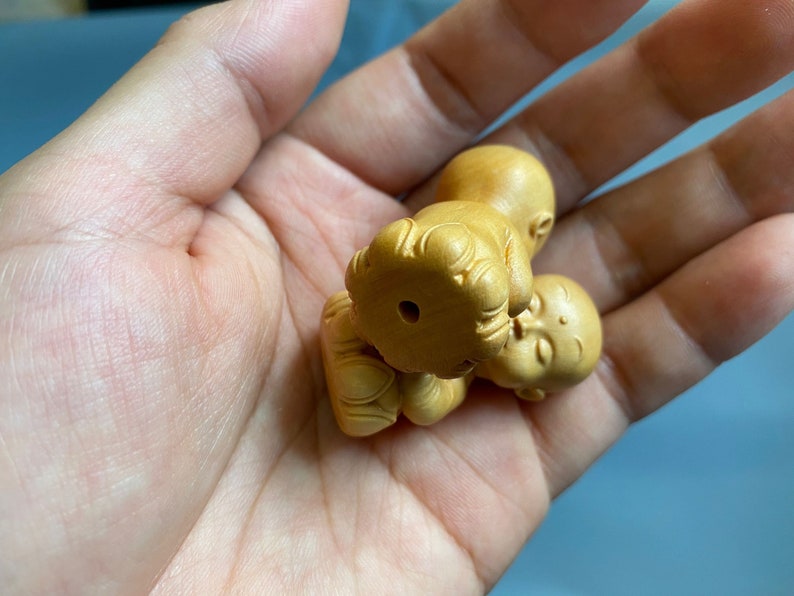 May include: A small, intricately carved wooden figurine of a baby Buddha. The figure is light brown in color and features detailed facial expressions and clothing. The Buddha is lying down, with its eyes closed, and is held in an open hand.