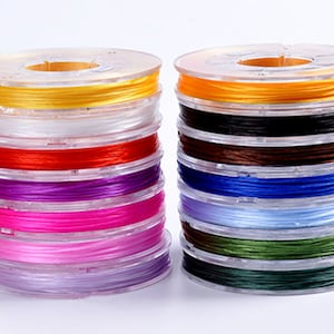 May include: Spools of colourful elastic string in various colours, including yellow, white, red, purple, pink, light pink, lavender, black, brown, blue, light blue, green, and dark green.