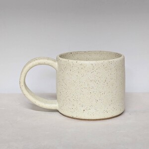 May include: A speckled ceramic mug with a rounded handle. The mug is off-white with small brown speckles throughout. The mug has a simple, cylindrical shape and a wide, curved handle.
