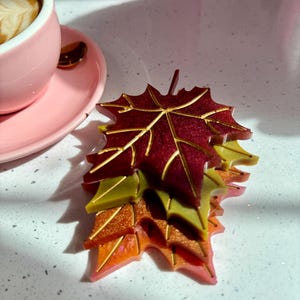 May include: A stack of four autumn-themed maple leaf-shaped coasters. The top coaster is a deep burgundy colour with gold veins. The next coaster is a light green with gold veins. The third coaster is a light orange with gold veins. The bottom coaster is a light pink with gold veins.