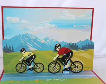 May include: A pop-up card featuring two cyclists riding bikes on a road with mountains in the background. The cyclists are wearing helmets and colorful clothing. The card is made of paper and has a 3D effect.