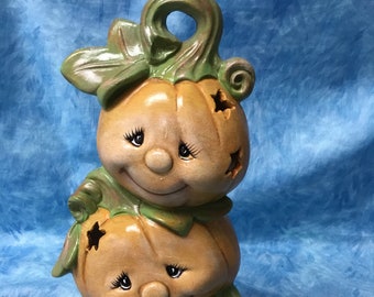 May include: A stack of three ceramic pumpkins with painted faces and star cutouts. The pumpkins are orange and brown with green stems and leaves.