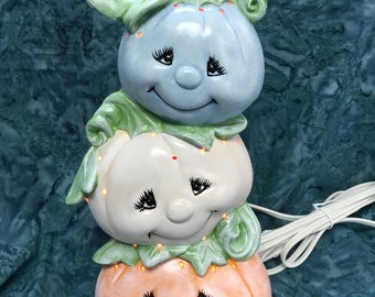 May include: A stack of three ceramic pumpkins with painted faces and glowing lights. The top pumpkin is blue, the middle pumpkin is white, and the bottom pumpkin is orange.