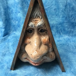 May include: A ceramic sculpture of a face peeking out from behind a triangular wooden frame. The face is painted with realistic details, including wrinkles and a large nose.