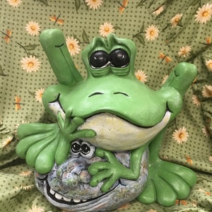 May include: A green frog figurine with large eyes and a wide smile, sitting on top of a smaller, gray frog figurine with a toothy grin. Both frogs are made of ceramic and have a whimsical, cartoonish style.
