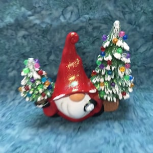 May include: A red and white gnome figurine holding two small Christmas trees decorated with colorful ornaments and white snow.
