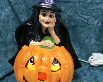 May include: A ceramic witch figurine with black hair and a purple hat sits on top of a lit jack-o'-lantern. The witch is holding her chin and looking to the side. The jack-o'-lantern has a wide smile and yellow eyes.