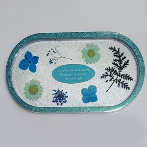May include: Oval-shaped resin tray with a teal border and a white glitter background. The tray features pressed flowers and leaves in shades of blue and green, along with a quote by Steven Wright: "If at first, you don't succeed, skydiving is not for you."