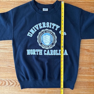 Op de afbeelding: Een marineblauw sweatshirt met de tekst "UNIVERSITY OF NORTH CAROLINA" in lichtblauw en wit. De sweatshirt heeft een ronde hals en lange mouwen. Een gele meetlint is verticaal geplaatst en meet ongeveer 71 cm.