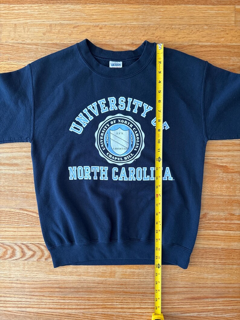 Op de afbeelding: Een marineblauw sweatshirt met de tekst "UNIVERSITY OF NORTH CAROLINA" in lichtblauw en wit. De sweatshirt heeft een ronde hals en lange mouwen. Een gele meetlint is verticaal geplaatst en meet ongeveer 71 cm.