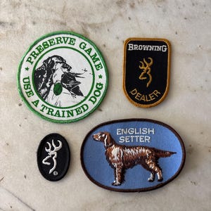 May include: Four embroidered patches: a round green-bordered patch with a dog and text "PRESERVE GAME USE A TRAINED DOG", a rectangular black Browning Dealer patch, a small oval black Browning logo patch, and an oval blue English Setter patch.