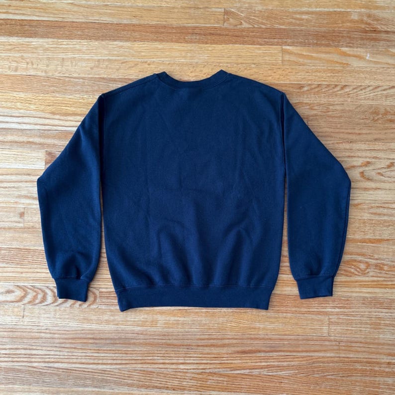 Op de afbeelding: Een marineblauw sweatshirt met ronde hals. De top met lange mouwen is gemaakt van een zacht materiaal en ligt plat op een houten oppervlak. De sweatshirt heeft een geribbelde kraag, manchetten en zoom.