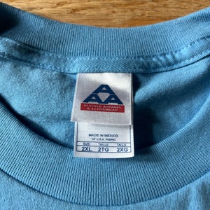 May include: A light blue t-shirt with a white tag that says "Alstyle Apparel & Activewear" and "Made in Mexico of U.S.A. Fabric". The tag also shows the size: 2XL, 2TG, 2XG.