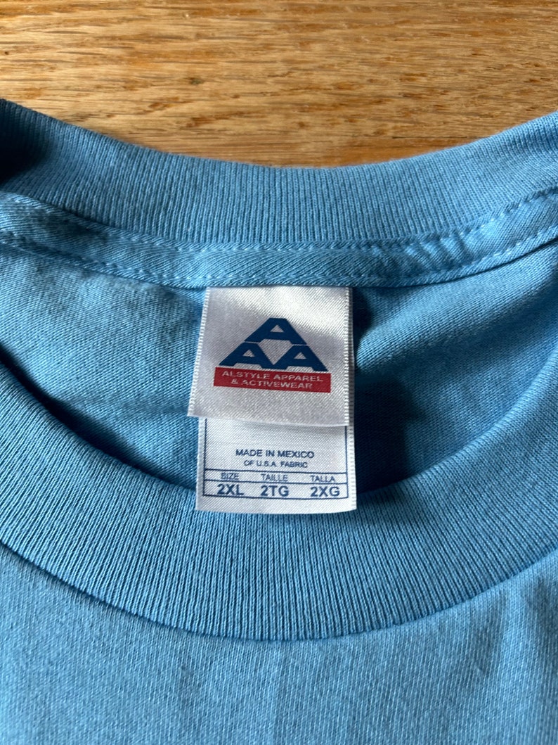 May include: A light blue t-shirt with a white tag that says "Alstyle Apparel & Activewear" and "Made in Mexico of U.S.A. Fabric". The tag also shows the size: 2XL, 2TG, 2XG.