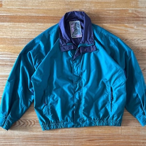 May include: Teal and purple vintage jacket with a button-up front, two side pockets, and a collared neckline. The jacket has a label on the collar. The jacket is made of a lightweight material, perfect for layering. The jacket is laid flat on a wooden surface.