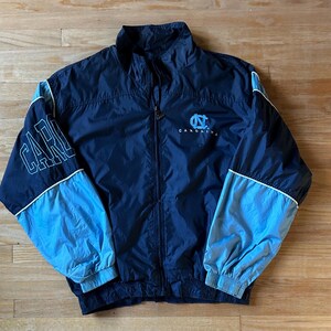May include: Navy blue and light blue zip-up jacket. The jacket features the word "CAROLINA" on the left sleeve and the "UNC" logo on the chest. White trim accents the sleeves and collar. The jacket is made of a lightweight material.