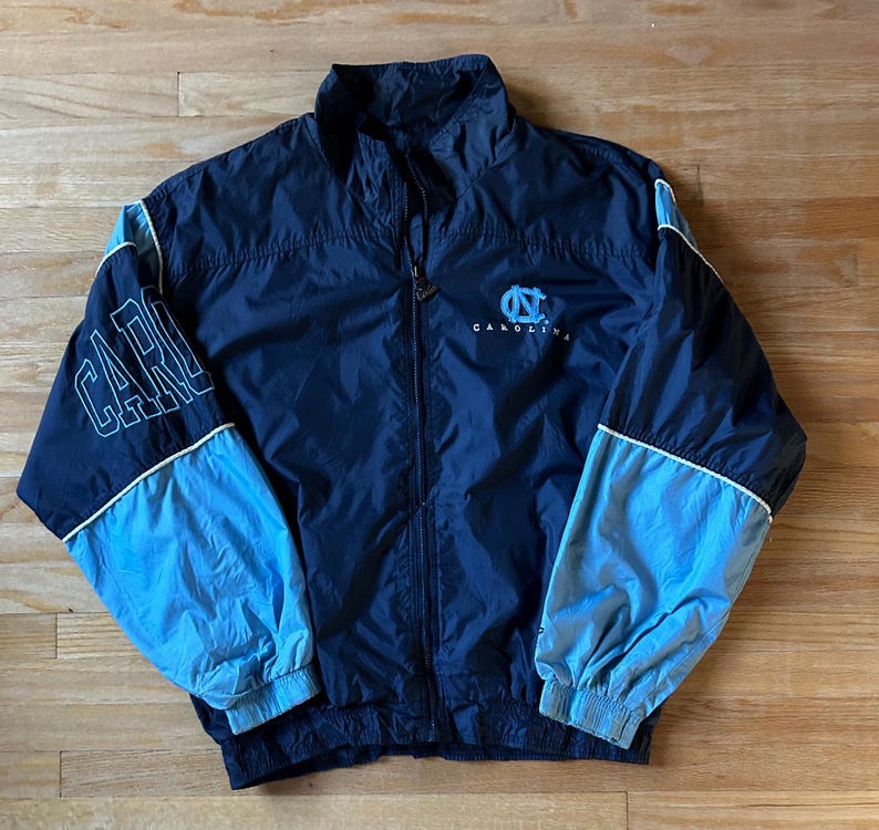 May include: Navy blue and light blue zip-up jacket. The jacket features the word "CAROLINA" on the left sleeve and the "UNC" logo on the chest. White trim accents the sleeves and collar. The jacket is made of a lightweight material.