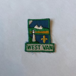 Könnte beinhalten: Ein gestickter Aufnäher in Grün und Blau mit einem weißen Leuchtturm, Bergen und einer Fleur-de-lis. Der Text "WEST VAN" ist in Weiß auf einem grünen Banner gestickt.