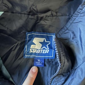 May include: Close-up of a navy blue Starter jacket with a blue and white logo tag. The tag reads "STARTER" and "XL MADE IN KOREA". The jacket has a zipper and a light blue lining.