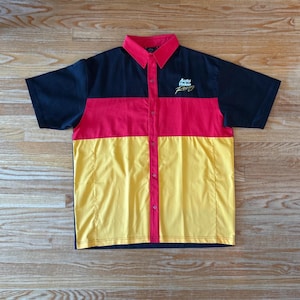 May include: Short-sleeved button-up shirt with a colour block design. The shirt features black sleeves and collar, a red centre panel, and a yellow lower panel. The text "Auto Value Racing" is embroidered on the chest.