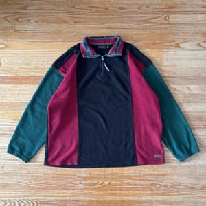 May include: A colour-blocked fleece pullover in navy, burgundy, and teal. The long-sleeved top has a quarter-zip closure and a collar with printed text. A small rectangular patch is visible near the bottom hem. The garment is laid flat on a wooden surface.
