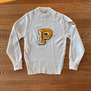 May include: Cream-colored vintage sweater with ribbed knit texture. Features a large yellow and black "P" patch with the word "SPEECH" vertically on the left side. A smaller yellow and black design is on the left sleeve. The sweater has a crew neck and long sleeves.