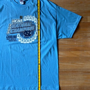 May include: A light blue t-shirt with a graphic of the 2005 NCAA National Champions, the Carolina Tar Heels basketball team. The graphic includes the team's logo and the text "NCAA 2005 National Champions Carolina Tar Heels. Basketball."