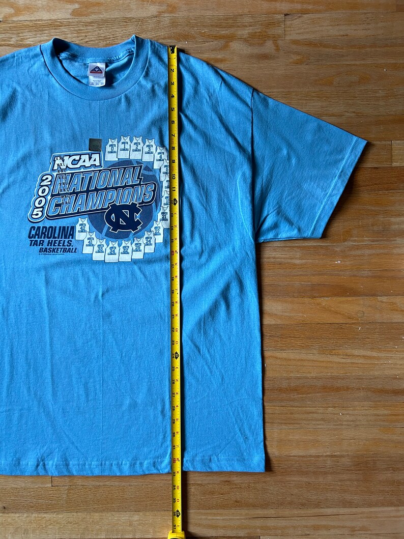 May include: A light blue t-shirt with a graphic of the 2005 NCAA National Champions, the Carolina Tar Heels basketball team. The graphic includes the team's logo and the text "NCAA 2005 National Champions Carolina Tar Heels. Basketball."