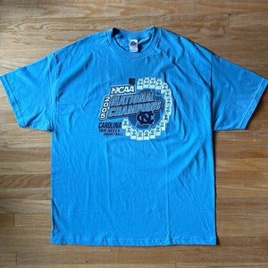 May include: A light blue t-shirt with a graphic of the 2005 NCAA National Champions, the Carolina Tar Heels basketball team. The graphic features the team's logo and a circle of basketball jerseys.
