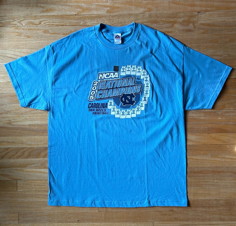 May include: A light blue t-shirt with a graphic of the 2005 NCAA National Champions, the Carolina Tar Heels basketball team. The graphic features the team's logo and a circle of basketball jerseys.