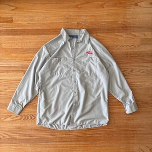 Può includere: Una camicia grigio chiaro con bottoni, maniche lunghe e due tasche sul petto. Il logo Coca-Cola è ricamato in rosso sul petto destro. La camicia è stesa su una superficie di legno.