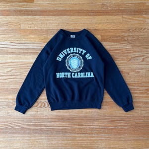 Op de afbeelding: Een marineblauw sweatshirt met de tekst "UNIVERSITY OF NORTH CAROLINA" in wit en lichtblauw. Het sweatshirt heeft een ronde hals en lange mouwen. Het embleem van de universiteit staat in het midden.
