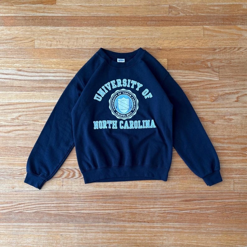 Op de afbeelding: Een marineblauw sweatshirt met de tekst "UNIVERSITY OF NORTH CAROLINA" in wit en lichtblauw. Het sweatshirt heeft een ronde hals en lange mouwen. Het embleem van de universiteit staat in het midden.
