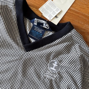 Op de afbeelding: Een zwart-wit pullover met pied-de-poule patroon en een marineblauwe bies rond de V-hals. De trui heeft een klein geborduurd logo en een aangehecht label. Het label luidt "Sunderland's Classic Pullover".