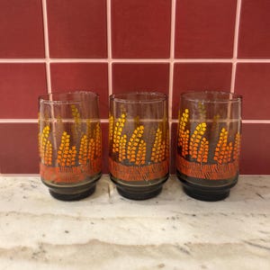 May include: Three vintage drinking glasses featuring a wheat-like pattern in orange and yellow. The design is set against a black background with an orange border. The glasses have a dark base and are displayed on a light-coloured surface.