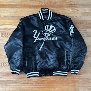 May include: Black satin Yankees baseball jacket with white accents on the collar, cuffs, and hem. The jacket features the team's logo and the word "Yankees" in white script across the front. The jacket has a button-up closure and two front pockets.