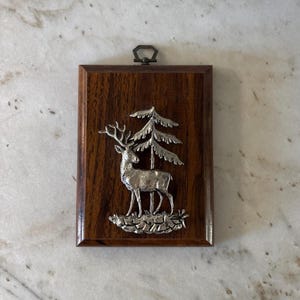 May include: A rectangular wooden plaque featuring a silver metal relief of a deer and a pine tree. The deer stands before the tree, and the plaque has a metal loop at the top for hanging. The wood has a dark brown finish.