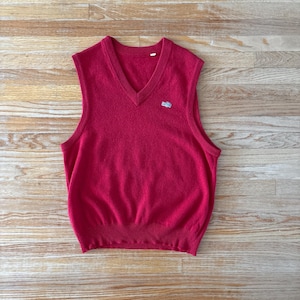 May include: A red V-neck sweater vest with a small embroidered emblem on the chest. The sleeveless garment is made of a soft, knitted material. The vest is a solid, vibrant red colour.