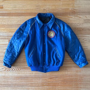 Könnte beinhalten: Blaue Collegejacke mit Lederärmeln und Knopfleiste. Die Jacke hat einen Aufnäher auf der Brust und gelbe Paspeln. Die Jacke ist aus Wolle und Leder.