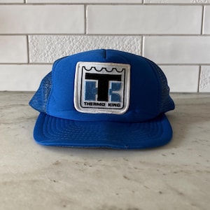 May include: A blue trucker hat with a mesh back and a curved brim. The front of the hat features a rectangular patch with the text "THERMO KING" and a stylized logo in black and blue.