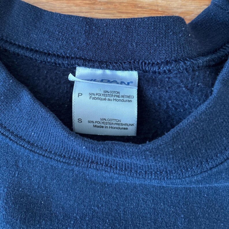 Op de afbeelding: Close-up van een marineblauw sweatshirt met een aangehecht label. Het label toont tekst in het Engels en Frans, met details over de samenstelling van de stof en de herkomst. De textuur van de stof van het sweatshirt is zichtbaar.