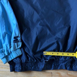 May include: A navy blue and light blue Starter jacket. The jacket has a drawstring waist and elastic cuffs. The Starter logo is visible on the light blue sleeve. The jacket is approximately 6 inches wide.