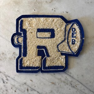 May include: A varsity-style letter patch featuring the letter R in a textured cream-coloured chenille fabric with a dark blue felt border. Attached to the right side of the letter is a small football shape with the letters DEB stacked vertically.