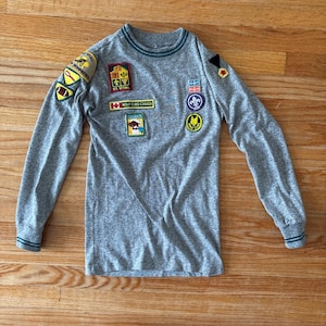 May include: A grey long-sleeved shirt adorned with several patches, including a Canadian flag and various badges. The shirt features green trim on the collar and cuffs. The patches display text and images, possibly related to a scouting or youth group.