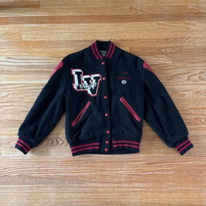 Peut inclure: Veste de style universitaire noire avec des détails rouges, affichant les lettres "LV" dans un design blanc et rouge sur la poitrine. La veste a des bordures rouges sur le col, les poignets et les poches, avec le chiffre "74" et le mot "forever" brodé.